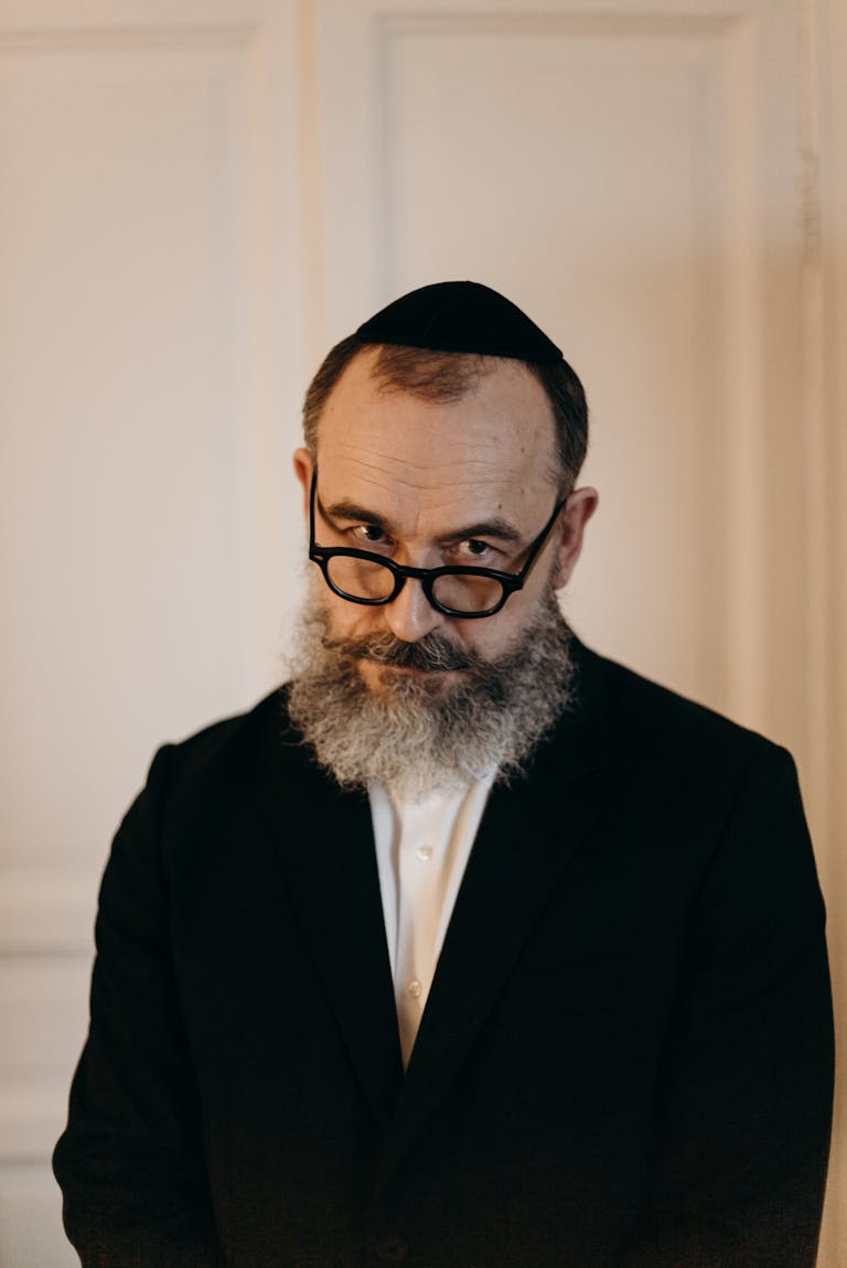 A bearded man wearing a kippah and glasses, dressed in a black suit, posing indoors.